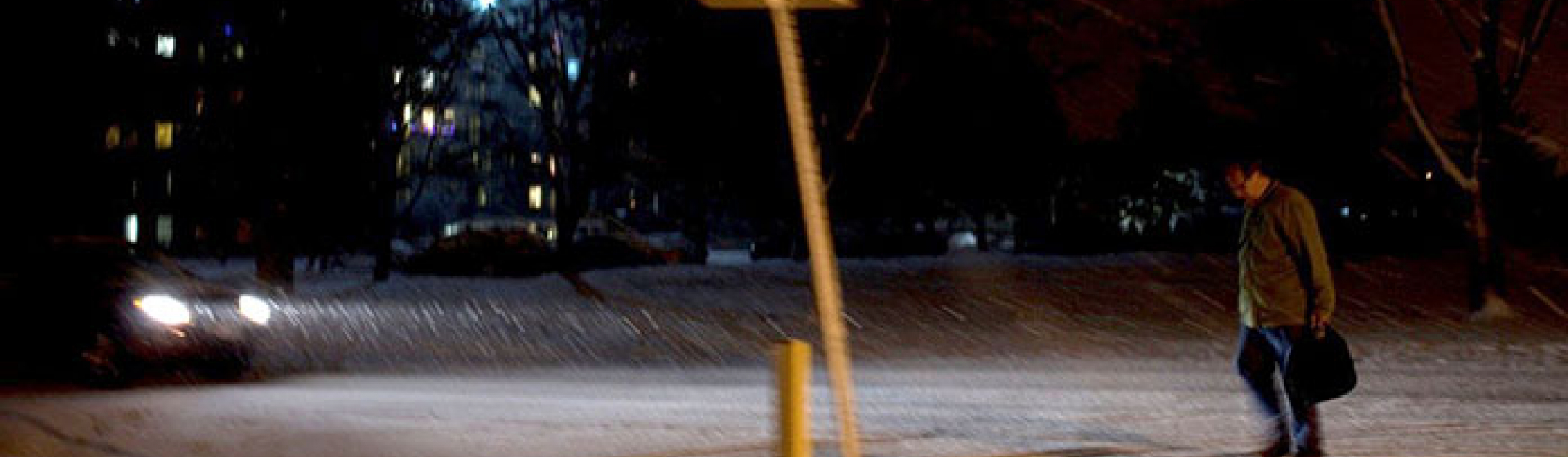 A man walks alone on a street at night with car headlights shining towards him